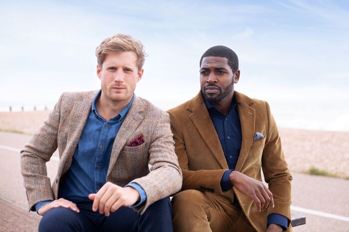 Two male models sitting wearing formal jackets with beach in the background