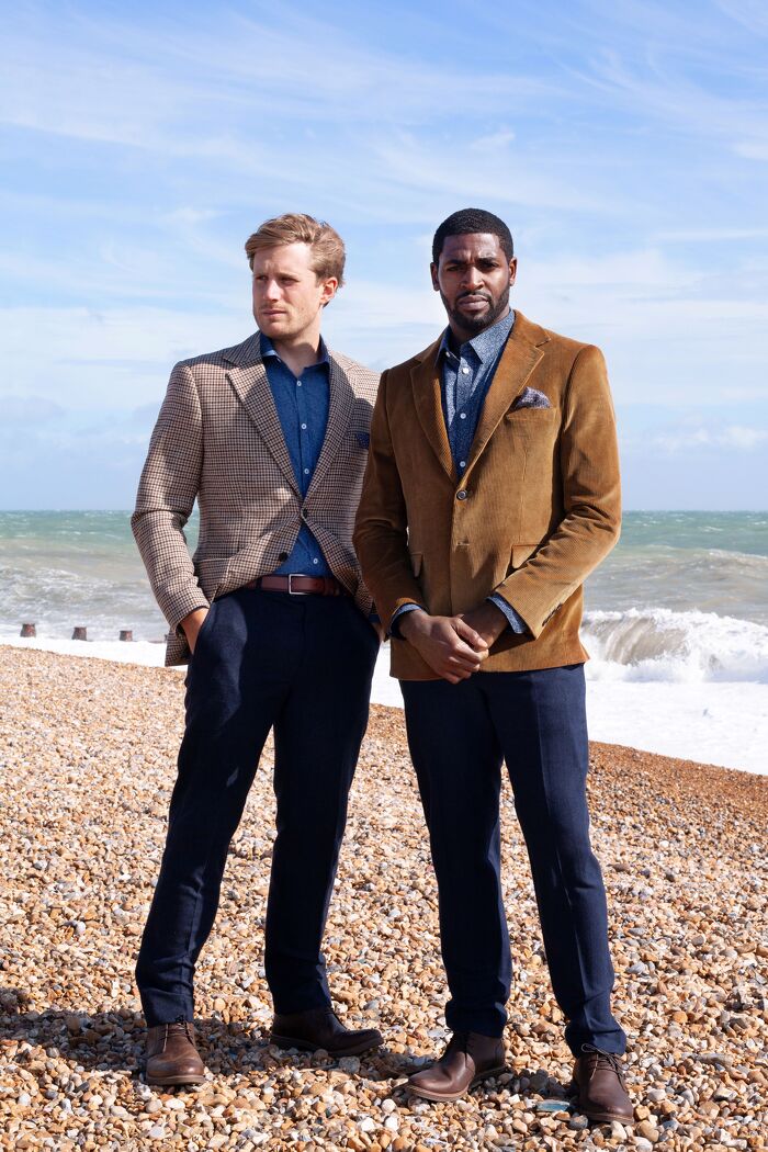 Two male models standing wearing formal jackets with beach in the background