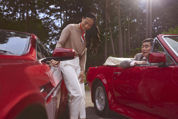 Woman leaning against red car talking to man in convertible car